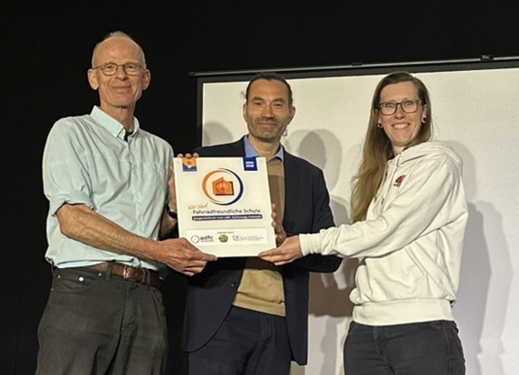 Nora Müller (links) vom ADFC SH übergibt die Plakette "Fahrradfreundliche Schule" an Mathias Kadow-Leder (Schulleiter Holstenschule) und Christian Wagner (Fahrradbeauftrager Holstenschule Neumünster)